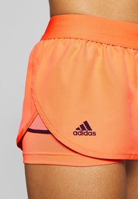 Pantalones cortos atléticos naranjas con cintura elástica, que presentan un diseño en capas y un logo púrpura de Adidas en la parte frontal. Textura de tela suave.