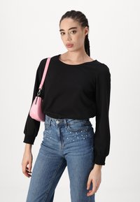 Svart langermet topp med elastiske mansjetter, kombinert med blå høyhelsesjeans med perleapplikasjoner. En liten rosa veske bæres over skulderen.