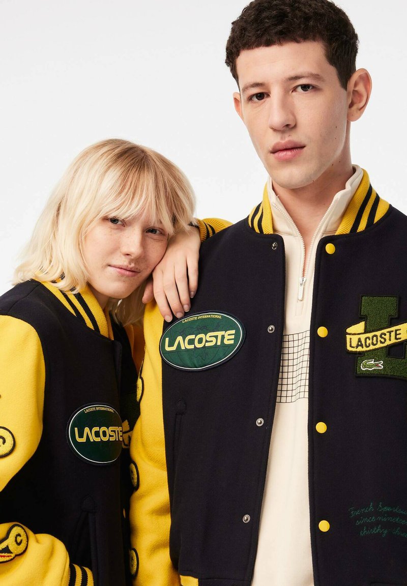 Vestes varsity avec un corps en laine noire et des manches jaunes, ornées de patches brodés en vert et blanc, col et poignets côtelés.