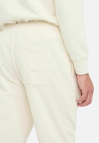 Pantalones de chándal color crema, hechos de una tela suave, con un bolsillo trasero que presenta un logo en relieve y una cintura elástica.
