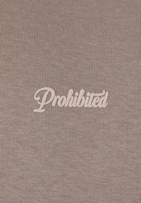 Világosbarna szövet texturált szövésű, amelyen fehér hímzett felirat található, ami így szól: "Tilos."