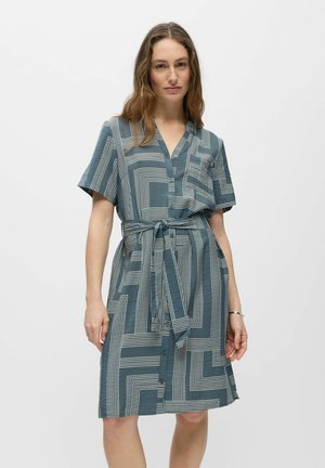 Femme portant une robe chemise manches courtes bleu-gris avec des lignes géométriques blanches, une ceinture à la taille et des boutons sur le devant, se tenant devant un fond blanc.