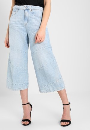 Person trägt hellblaue, weit geschnittene, verkürzte Jeans und schwarze, hochhackige Sandalen mit Knöchelriemen und steht vor einem einfarbigen Hintergrund.