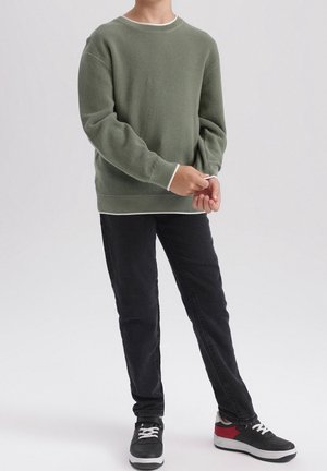 Osoba ubrana w oliwkowy sweter o fakturze, czarne dżinsy oraz czarne trampki z białymi sznurowadłami i czerwonymi akcentami, stojąca na jasnym, jednolitym tle.