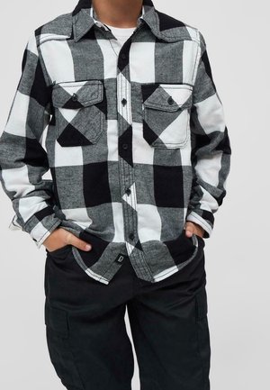 Osoba ubrana w czarno-białą koszulę w kratę z dwoma kieszeniami na piersi i czarne spodnie bojówki, ręce w kieszeniach.