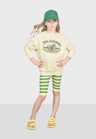 Felpa beige con design stampato verde, leggings a righe verdi e gialle, ciabatte gialle con testo nero e berretto da baseball verde.