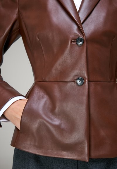Gros plan d'une personne portant un blazer en cuir marron avec deux boutons noirs, une main dans la poche, et un poignet de chemise blanc visible.