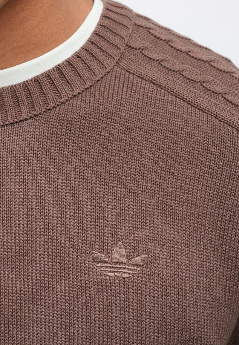 Brown knitted sweater featuring a ribbed crew neckline and textured detailing on shoulders. Includes an embroidered logo on the chest.