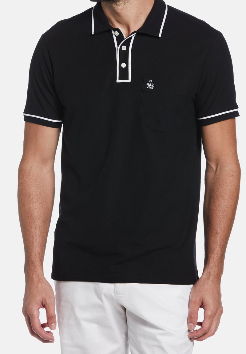 Homme portant un polo noir à manches courtes avec un bord blanc et un petit logo sur la poche, associé à un pantalon blanc.