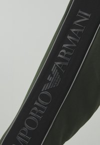 Olive zelená taška s výrazným černým popruhem s raženým logem "Emporio Armani" a stříbrnými akcenty podél okrajů.