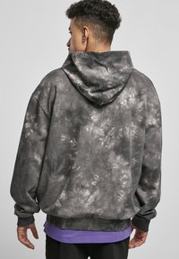 Hoodie s crno-bijelim tie-dye uzorkom, opuštenog kroja i rebrastim rukavima, složen preko ljubičaste majice.