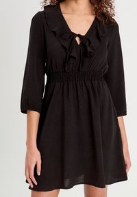 Femme portant une robe noire à manches longues avec un décolleté en V à volants et un noeud, une taille élastique et une jupe évasée.