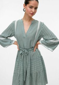 Vestido verde con estampado, con escote en V, mangas largas y cintura ajustada. Presenta motivos geométricos y de hojas con texturas alternadas.