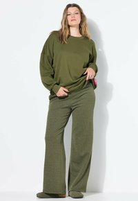 Vrouw in een olijfgroen oversized sweatshirt, met een patroon op wijde broekspijpen en bijpassende schoenen, staand tegen een witte achtergrond.