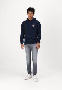 Felpa blu navy con tasca frontale ed emblema, abbinata a jeans grigio chiaro e sneakers bianche, in piedi davanti a uno sfondo neutro.