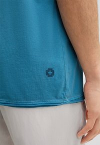 T-shirt en coton bleu avec une texture douce, arborant un petit logo bleu foncé sur le côté inférieur et une coupe décontractée. Coutures visibles.