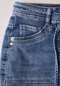Tissu en denim bleu clair avec un design classique à cinq poches. Présente des coutures orange, des rivets en métal et une texture lisse.