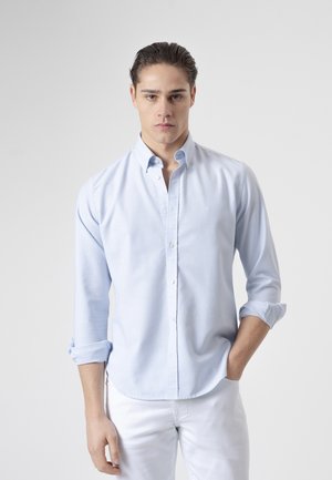 Joven con el cabello peinado hacia atrás, vistiendo una camisa azul claro abotonada con las mangas remangadas y pantalones blancos, de pie frente a un fondo liso.