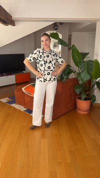 Femme les mains sur les hanches, portant une chemise fleurie noire et blanche, pantalon blanc, debout sur un sol en bois à côté d'un canapé orange et d'une grande plante en pot.