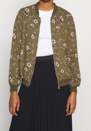 Person wearing an olive green floral zip-up bomber jacket, white top, and black pleated skirt, standing with arms relaxed.