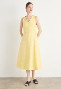 Rochie midi din in galben, cu decolteu în V și design fără mâneci. Are un corset ajustat și o fustă fluidă. Mule negre cu vârful deschis.