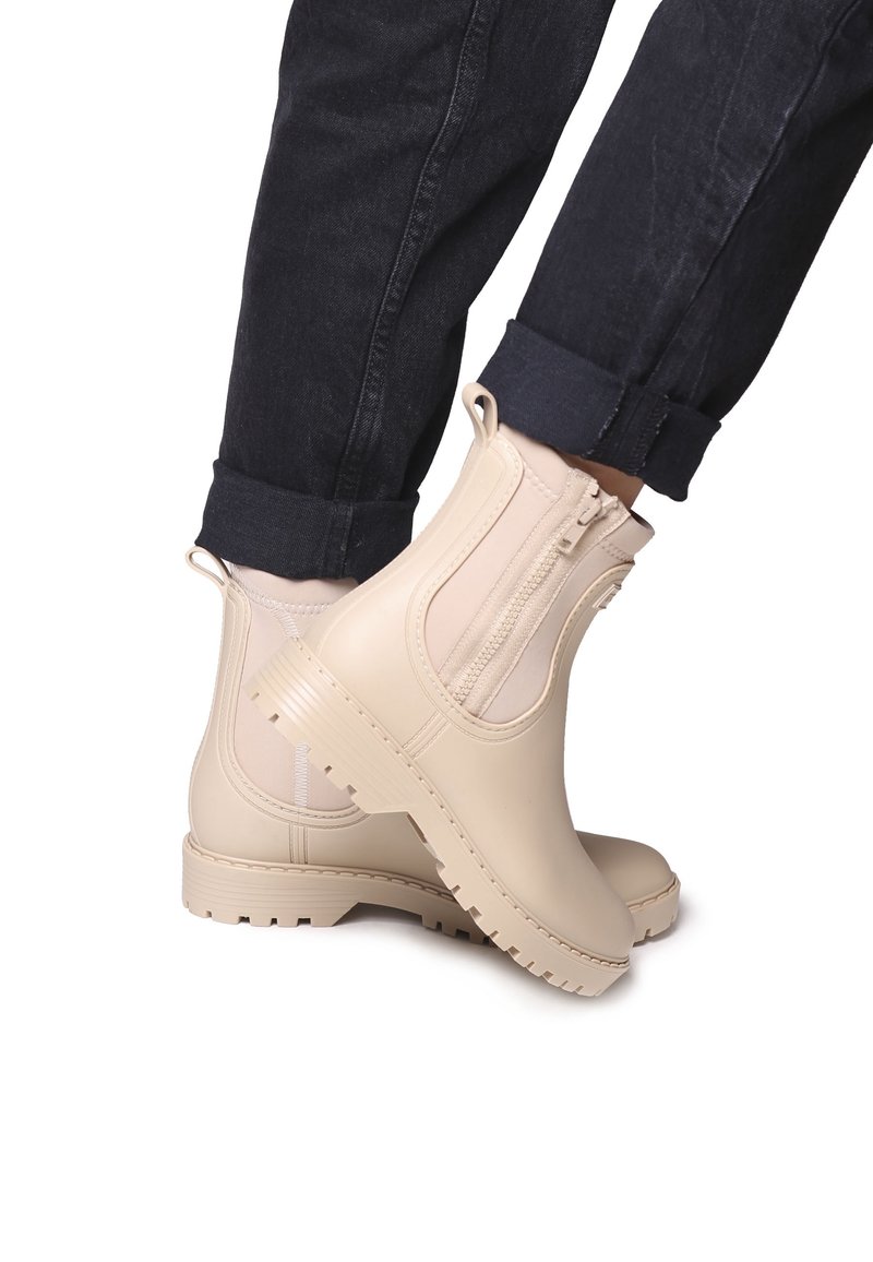 Bottes beiges à la cheville avec fermeture éclair latérale et languettes, portées avec un jean foncé roulé, vues de profil sur fond blanc.