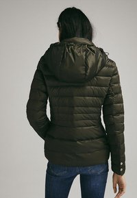 Veste matelassée vert foncé avec capuche, dotée de surpiqûres horizontales et de boutons-pression aux poignets. Texture lisse et rembourrée.