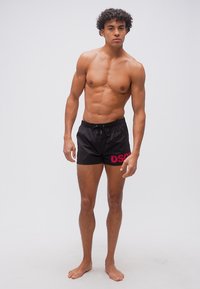Jeune homme aux cheveux bouclés debout pieds nus, portant un short de bain noir avec les lettres rouges "DSQ" sur un fond clair uni.