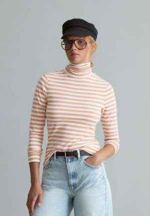 Vrouw die een beige-wit gestreepte coltrui draagt, lichtblauwe spijkerbroek, zwarte riem, grote bril en een zwarte platte pet, staand met één hand in de zak.