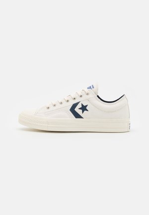 Biały niski sneaker z granatowym logo w kształcie gwiazdy chevron, sznurowanym wzorem i płaską gumową podeszwą na białym tle.