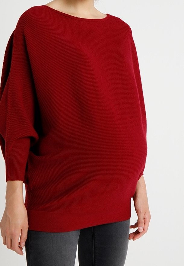 Punainen ribbineulepullover, jossa on leveä kaula-aukko ja rento istuvuus. Tässä neuleessa on pudotetut olkapäät ja pitkät hihat, ja se on yhdistetty tummanharmaisiin farkkuihin.