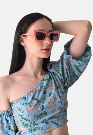 Rosa Cat-Eye-Sonnenbrille mit dunklen Gläsern. Das Modell trägt ein hellblaues Oberteil mit floralem Muster in Grün und Orange. Glattes Material, einseitiges Design.