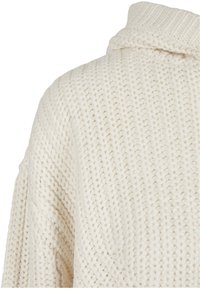 Kremfarget strikket genser med høy krage. Har en tykk tekstur og løs, avslappet passform, med ribbed mønstre på overflaten.