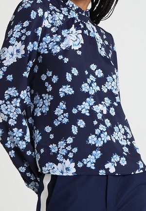 Marineblauwe blouse met lange mouwen en een lichtblauw-wit bloemenpatroon, gecombineerd met marineblauwe broek met een witte zijstreep.