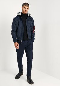 Chaqueta de bombardero navy con capucha gris, puños acanalados, cierre de cremallera en el frente y bolsillos laterales. Combinada con pantalones oscuros y botas negras hasta el tobillo.