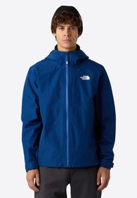 Mladý muž v modré nepromokavé bundě The North Face s kapucí a černou čepicí, stojící na jednotném pozadí.