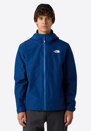 Młody mężczyzna ubrany w niebieską wodoodporną kurtkę The North Face z kapturem i czarną czapką beanie, stojący na jednolitym tle.