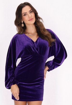 Woman with long wavy hair wears purple velvet dress with long puffed sleeves, gold chain necklace, and gold statement earrings against white background.