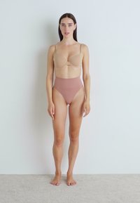 Beige underwire-bh med formede kopper og justerbare stropper, kombinert med høyt midje, sømløse rosa truser. Modellen står barføtt mot en ensfarget vegg.