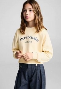 Felpa gialla con testo "Mer et étoiles" in blu navy, polsini a costine, indossata con pantaloni alti blu scuro con bottoni argentati.