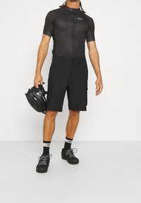 Mies, který má na sobě černý cyklistický dres a šortky, drží černou cyklistickou helmu Fox, nosí černé ponožky a černé cyklistické boty.