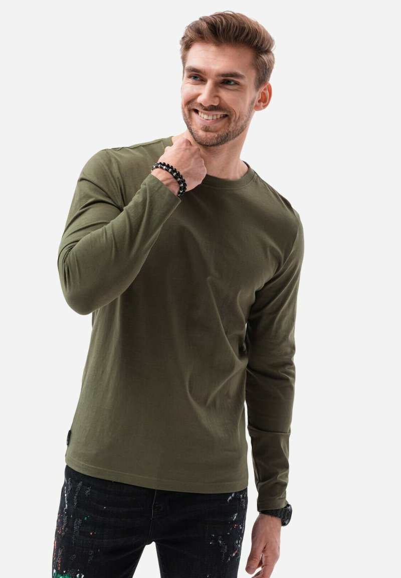 Camicia verde a maniche lunghe realizzata in morbido cotone, con scollatura rotonda e discreti dettagli di cucitura. Indossata con jeans scuri e un braccialetto.