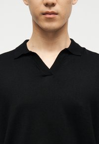 Jeune personne portant un pull noir à col en V avec un petit col, visible du cou jusqu'à la partie supérieure de la poitrine sur un fond uni.