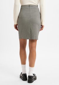 Rock mit Hahnentrittmuster und enger Silhouette, mit einem Reißverschluss auf der Rückseite und einer Schlitzeinschaffung. Getragen mit schwarzen Slippern und weißen Socken.