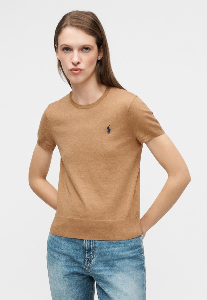 Pull marron à manches courtes avec un col rond côtelé, arborant un petit logo bleu marine sur la poitrine et un ourlet ajusté. Porté avec un jeans en denim bleu.