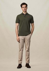 Tmavě zelené polokošile s krátkým rukávem, zdobené námořnickým límečkem a vyšitým červeným logem, v kombinaci s béžovými cargo kalhotami a hnědými botami.