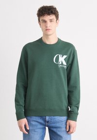 Hanorac verde din bumbac cu manșete și guler din material ribbed. Prezintă un mare logo alb "CK" împreună cu text. Purtați cu blugi albaștri.