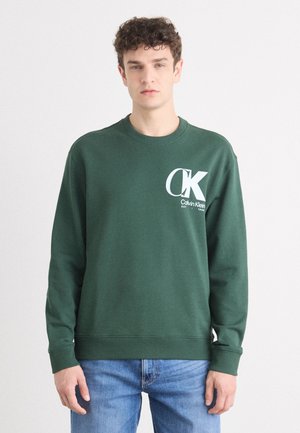 Zelená bavlněná mikina s žebrovanými manžetami a výstřihem. Obsahuje velké bílé logo "CK" s textem. Nosená s modrými džíny.
