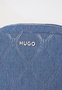 Pochette in denim con un motivo a zigzag trapuntato, caratterizzata da un logo 'HUGO' in argento e cuciture a contrasto su uno sfondo di tessuto blu.