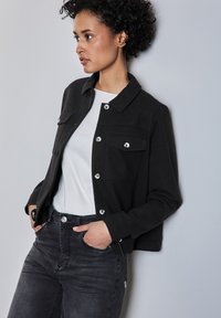 Veste noire courte avec boutons argentés, deux poches avant et un col de chemise. Portée sur une chemise blanche avec un jean noir taille haute.